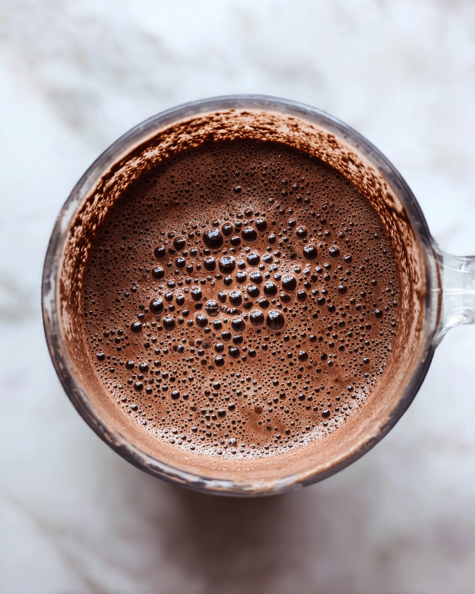 This image shows a close-up top view inside a blender jar filled with a thick chocolate-colored liquid. The liquid has many small bubbles on its surface, giving it a slightly frothy texture. The sides of the blender jar are coated with a thin layer of the chocolate mixture, adding a smooth and creamy look to the image. The jar itself is clear and the photo is set against a white marbled textured background. photo taken with an iphone --ar 4:5 --v 7